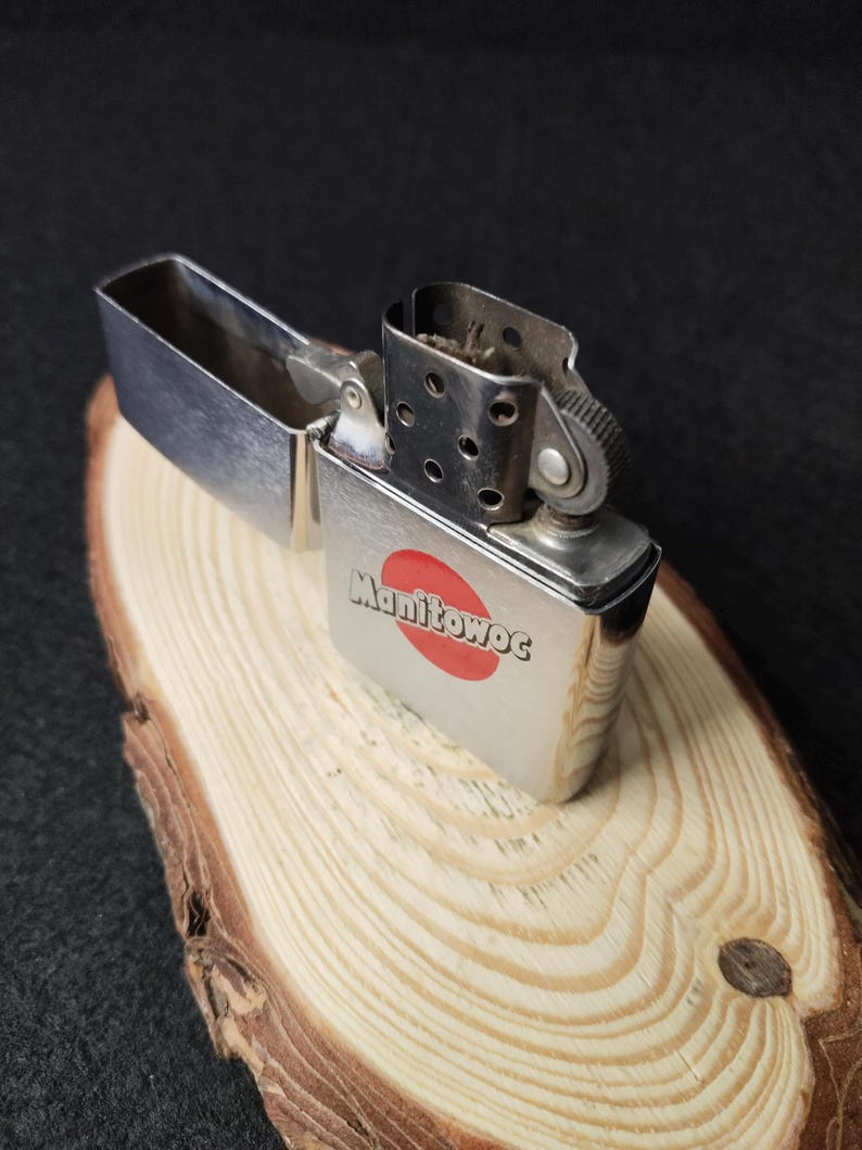 Peut inclure: Un briquet argent&eacute; ouvert, avec le mot "Manitowoc" et un cercle rouge imprim&eacute;s. Il est pos&eacute; sur une surface en bois clair, sur fond sombre.