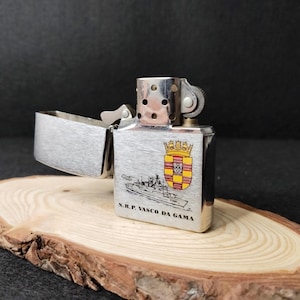 Może przedstawiać: Srebrna metalowa zapalniczka z otwartą g&oacute;rą. Posiada ilustrację herbu i statku oraz napis "N.R.P. VASCO DA GAMA".