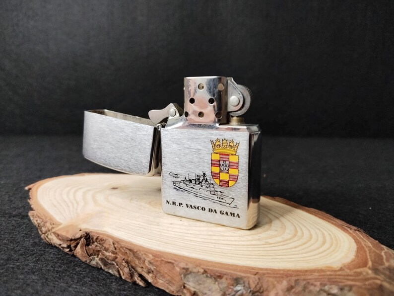 Może przedstawiać: Srebrna metalowa zapalniczka z otwartą g&oacute;rą. Posiada ilustrację herbu i statku oraz napis "N.R.P. VASCO DA GAMA".