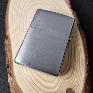 Peut inclure: Un briquet rectangulaire argent&eacute; avec une finition en m&eacute;tal bross&eacute;. Il est pos&eacute; sur une surface en bois circulaire brun clair avec des motifs de grain visibles. Le fond est sombre.
