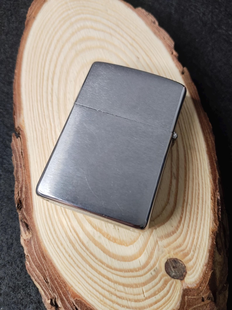 Peut inclure: Un briquet rectangulaire argent&eacute; avec une finition en m&eacute;tal bross&eacute;. Il est pos&eacute; sur une surface en bois circulaire brun clair avec des motifs de grain visibles. Le fond est sombre.