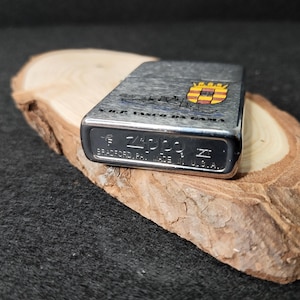 Może przedstawiać: Srebrna zapalniczka Zippo z teksturowanym wykończeniem, z napisem "V.R.F. VASCO DA GAMA" i herbem z czerwonymi, ż&oacute;łtymi i niebieskimi detalami. Zapalniczka jest wyprodukowana w USA i spoczywa na kawałku drewna.