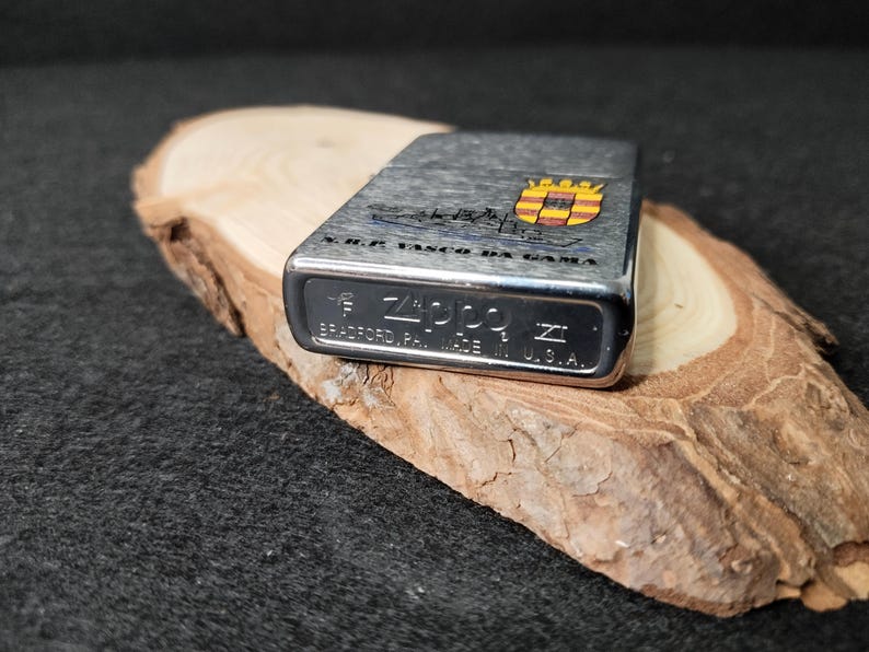 Może przedstawiać: Srebrna zapalniczka Zippo z teksturowanym wykończeniem, z napisem "V.R.F. VASCO DA GAMA" i herbem z czerwonymi, ż&oacute;łtymi i niebieskimi detalami. Zapalniczka jest wyprodukowana w USA i spoczywa na kawałku drewna.