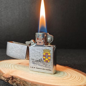 Może przedstawiać: Srebrna zapalniczka z płomieniem, z ilustracją statku i napisem "N.R.P. VASCO DA GAMA". Zapalniczka jest otwarta, odsłaniając wnętrze, i spoczywa na drewnianej powierzchni.