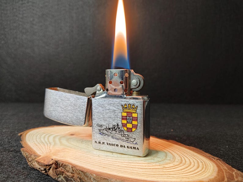 Może przedstawiać: Srebrna zapalniczka z płomieniem, z ilustracją statku i napisem "N.R.P. VASCO DA GAMA". Zapalniczka jest otwarta, odsłaniając wnętrze, i spoczywa na drewnianej powierzchni.