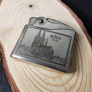 May include: A silver-toned lighter with a detailed engraving of the Cologne Cathedral, featuring the text "KÖLN-DOM". The lighter is rectangular with a hinged top and rests on a wooden surface.