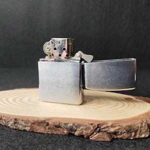 Peut inclure: Un briquet argent&eacute; ouvert, r&eacute;v&eacute;lant l'int&eacute;rieur, pos&eacute; sur une tranche de bois. Le briquet a un corps rectangulaire et un couvercle &agrave; charni&egrave;re. La tranche de bois pr&eacute;sente des cernes de croissance et des bords d'&eacute;corce. Fond sombre.