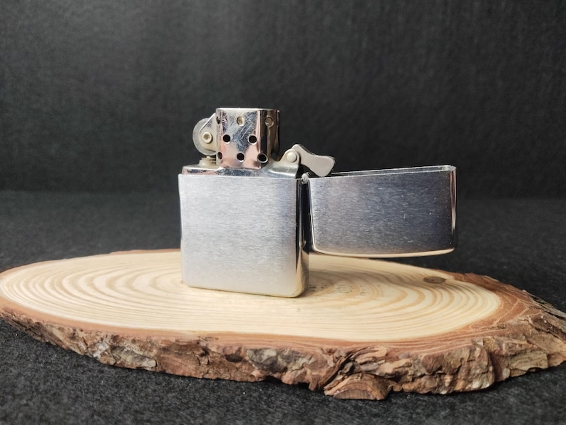 Peut inclure: Un briquet argent&eacute; ouvert, r&eacute;v&eacute;lant l'int&eacute;rieur, pos&eacute; sur une tranche de bois. Le briquet a un corps rectangulaire et un couvercle &agrave; charni&egrave;re. La tranche de bois pr&eacute;sente des cernes de croissance et des bords d'&eacute;corce. Fond sombre.