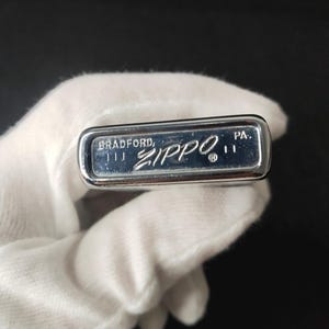 Peut inclure: Un briquet Zippo argent&eacute; avec l'inscription "BRADFORD, ZIPPO, PA." grav&eacute;e en bas. Le briquet est tenu par une main gant&eacute;e sur un fond sombre. Le briquet est rectangulaire et a une surface brillante et r&eacute;fl&eacute;chissante.
