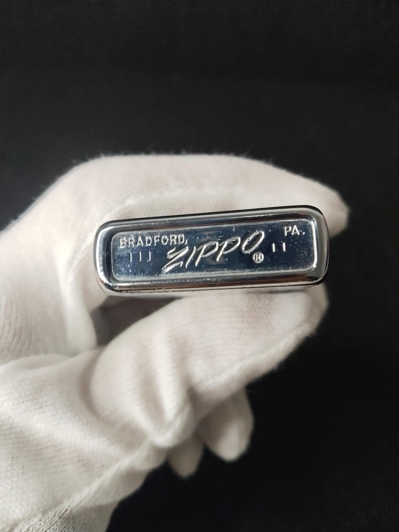 Peut inclure: Un briquet Zippo argent&eacute; avec l'inscription "BRADFORD, ZIPPO, PA." grav&eacute;e en bas. Le briquet est tenu par une main gant&eacute;e sur un fond sombre. Le briquet est rectangulaire et a une surface brillante et r&eacute;fl&eacute;chissante.