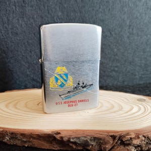 Puede incluir: Encendedor de metal plateado con un dise&ntilde;o de barco y el texto "USS JOSEPHUS DANIELS OLG-27". El encendedor tiene forma rectangular y una tapa con bisagras. El dise&ntilde;o incluye un escudo con detalles azules y amarillos.