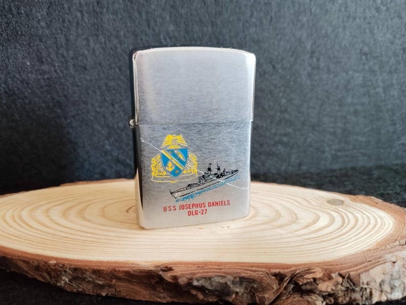 Puede incluir: Encendedor de metal plateado con un dise&ntilde;o de barco y el texto "USS JOSEPHUS DANIELS OLG-27". El encendedor tiene forma rectangular y una tapa con bisagras. El dise&ntilde;o incluye un escudo con detalles azules y amarillos.