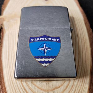 Może przedstawiać: Srebrny metalowy zapalniczka z niebiesko-białym emblematem. Emblemat przedstawia gwiazdę wewnątrz kompasu, tekst "STANAVFORLANT" i falisty wz&oacute;r na dole. Zapalniczka jest prostokątna z zaokrąglonymi krawędziami.