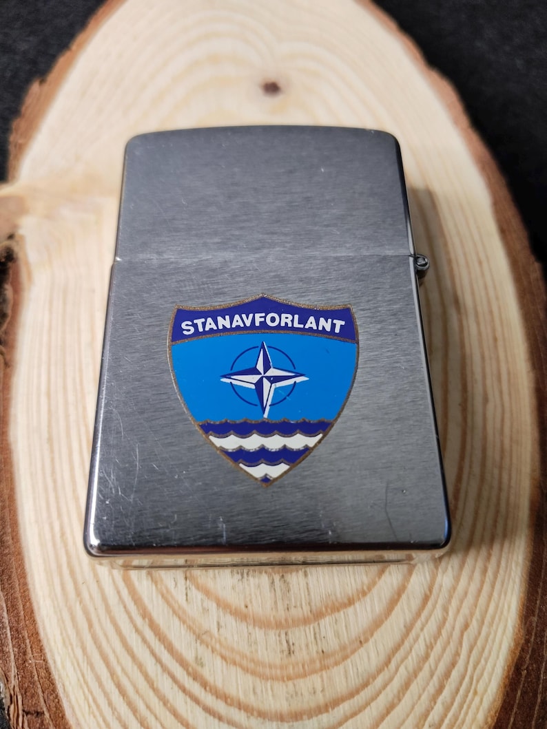 Może przedstawiać: Srebrny metalowy zapalniczka z niebiesko-białym emblematem. Emblemat przedstawia gwiazdę wewnątrz kompasu, tekst "STANAVFORLANT" i falisty wz&oacute;r na dole. Zapalniczka jest prostokątna z zaokrąglonymi krawędziami.