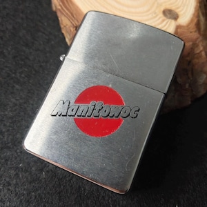 Peut inclure: Un briquet argent&eacute; avec un cercle rouge et le mot "Manitowoc" imprim&eacute; dessus. Le briquet rectangulaire a un couvercle &agrave; charni&egrave;re. L'arri&egrave;re-plan est une surface en bois.