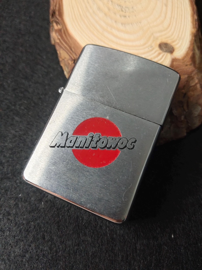 Peut inclure: Un briquet argent&eacute; avec un cercle rouge et le mot "Manitowoc" imprim&eacute; dessus. Le briquet rectangulaire a un couvercle &agrave; charni&egrave;re. L'arri&egrave;re-plan est une surface en bois.