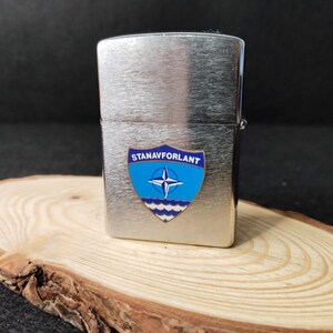 Może przedstawiać: Srebrna zapalniczka z niebiesko-białą odznaką z napisem "STANAVFORLANT". Odznaka przedstawia r&oacute;żę wiatr&oacute;w i fale. Zapalniczka spoczywa na drewnianym plastrze.