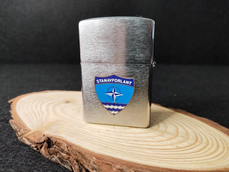 Może przedstawiać: Srebrna zapalniczka z niebiesko-białą odznaką z napisem "STANAVFORLANT". Odznaka przedstawia r&oacute;żę wiatr&oacute;w i fale. Zapalniczka spoczywa na drewnianym plastrze.