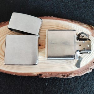 Puede incluir: Tres encendedores plateados se exhiben sobre una superficie de madera. Uno est&aacute; abierto, revelando su interior, mientras que los otros dos est&aacute;n cerrados. Los encendedores tienen forma rectangular con bordes redondeados y un acabado met&aacute;lico cepillado.