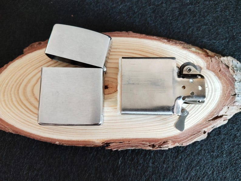 Puede incluir: Tres encendedores plateados se exhiben sobre una superficie de madera. Uno est&aacute; abierto, revelando su interior, mientras que los otros dos est&aacute;n cerrados. Los encendedores tienen forma rectangular con bordes redondeados y un acabado met&aacute;lico cepillado.