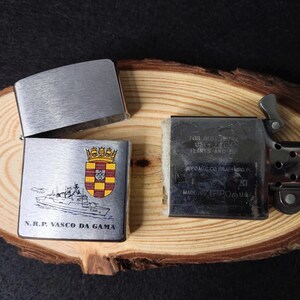 Może przedstawiać: Srebrna metalowa zapalniczka z otwartą pokrywą, odsłaniająca wnętrze. Zapalniczka posiada herb i ilustrację statku z napisem "N.R.P. VASCO DA GAMA". Zapalniczka leży na drewnianej powierzchni.