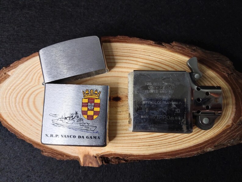Może przedstawiać: Srebrna metalowa zapalniczka z otwartą pokrywą, odsłaniająca wnętrze. Zapalniczka posiada herb i ilustrację statku z napisem "N.R.P. VASCO DA GAMA". Zapalniczka leży na drewnianej powierzchni.