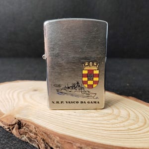 Może przedstawiać: Srebrny metalowy zapalniczka z ilustracją statku i napisem "N.R.P. VASCO DA GAMA". Zapalniczka posiada herb z koroną i tarczą z czerwonymi, ż&oacute;łtymi i czarnymi detalami. Zapalniczka jest prezentowana na drewnianej powierzchni.