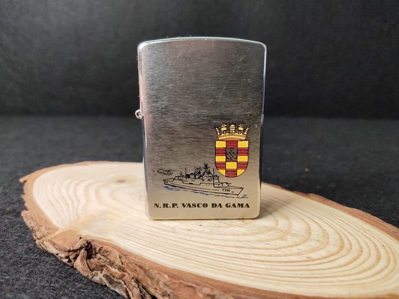 Może przedstawiać: Srebrny metalowy zapalniczka z ilustracją statku i napisem "N.R.P. VASCO DA GAMA". Zapalniczka posiada herb z koroną i tarczą z czerwonymi, ż&oacute;łtymi i czarnymi detalami. Zapalniczka jest prezentowana na drewnianej powierzchni.
