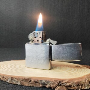 Peut inclure: Un briquet en m&eacute;tal argent&eacute; avec une flamme allum&eacute;e, sur fond sombre. Le briquet est ouvert, r&eacute;v&eacute;lant la flamme. Il repose sur une tranche de bois avec un bord naturel.