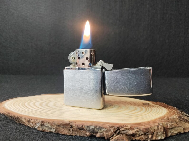 Peut inclure: Un briquet en m&eacute;tal argent&eacute; avec une flamme allum&eacute;e, sur fond sombre. Le briquet est ouvert, r&eacute;v&eacute;lant la flamme. Il repose sur une tranche de bois avec un bord naturel.