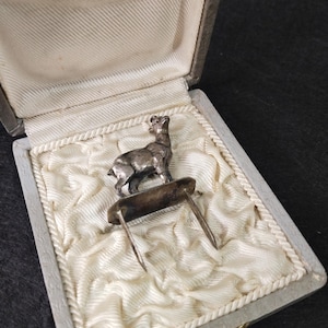 Puede incluir: Un alfiler de metal plateado con una pequeña figura de cabra de pie unida a una base rectangular con dos puntas largas y delgadas que se extienden hacia abajo, presentado dentro de una caja de exhibición articulada forrada de tela clara.