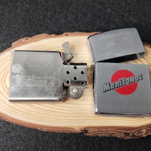 Peut inclure: Un briquet Zippo argent&eacute; avec le couvercle ouvert, r&eacute;v&eacute;lant l'int&eacute;rieur. Le briquet porte le mot "Manitowoc" en blanc sur un cercle rouge. Il est pos&eacute; sur une surface en bois.