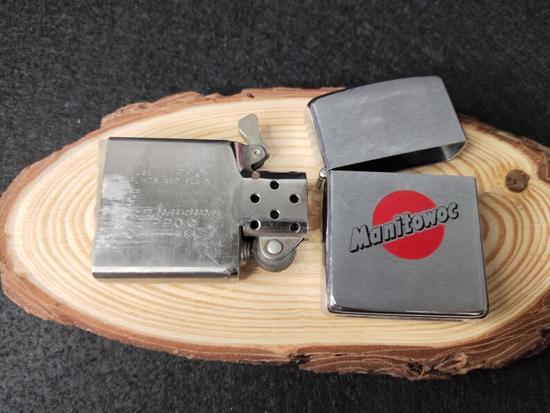 Peut inclure: Un briquet Zippo argent&eacute; avec le couvercle ouvert, r&eacute;v&eacute;lant l'int&eacute;rieur. Le briquet porte le mot "Manitowoc" en blanc sur un cercle rouge. Il est pos&eacute; sur une surface en bois.