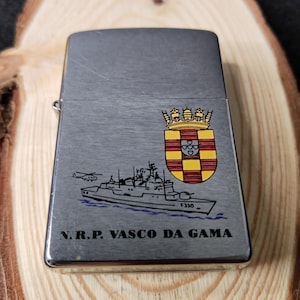 Może przedstawiać: Srebrna zapalniczka z klapką na zawiasach. Zapalniczka posiada ilustrację statku, helikoptera i herbu. Tekst "N.R.P. VASCO DA GAMA" jest wydrukowany pod ilustracją statku.