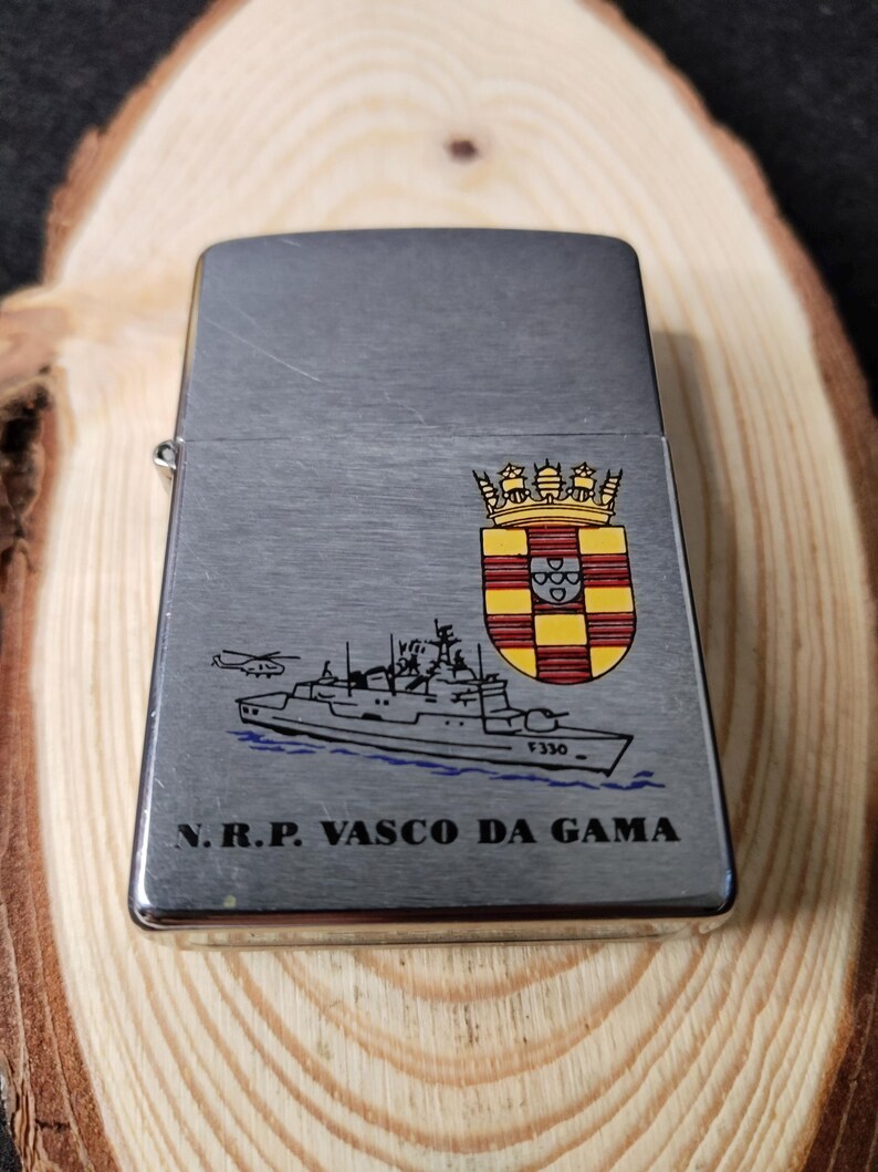 Może przedstawiać: Srebrna zapalniczka z klapką na zawiasach. Zapalniczka posiada ilustrację statku, helikoptera i herbu. Tekst "N.R.P. VASCO DA GAMA" jest wydrukowany pod ilustracją statku.