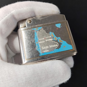 May include: A silver-toned lighter with a map of Saudi Arabia and surrounding areas. The map includes cities like Cairo, Tehran, and Aden. The lighter is rectangular with a hinged top.