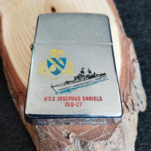 Puede incluir: Un encendedor plateado con una ilustraci&oacute;n detallada de un barco y un escudo. El texto "USS JOSEPHUS DANIELS DLG-27" est&aacute; impreso debajo. El encendedor est&aacute; sobre un trozo de madera.