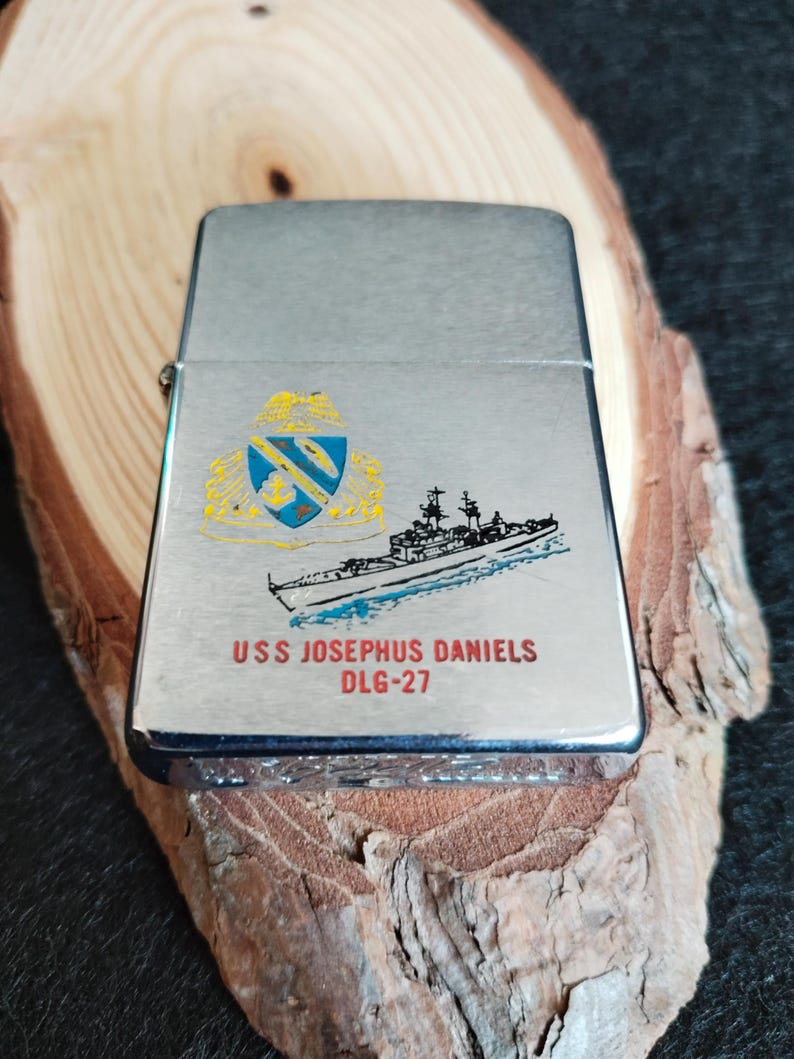 Puede incluir: Un encendedor plateado con una ilustraci&oacute;n detallada de un barco y un escudo. El texto "USS JOSEPHUS DANIELS DLG-27" est&aacute; impreso debajo. El encendedor est&aacute; sobre un trozo de madera.