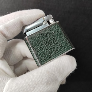 Puede incluir: Un encendedor vintage de forma rectangular. El encendedor tiene una parte superior de color plateado y un exterior texturizado de color verde oscuro similar al cuero. El encendedor se sostiene con un guante blanco.