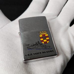 Może przedstawiać: Srebrna metalowa zapalniczka ze szczotkowanym wykończeniem. Zapalniczka posiada kolorowy emblemat i ilustrację statku z napisem "N.R.P. VASCO DA GAMA".
