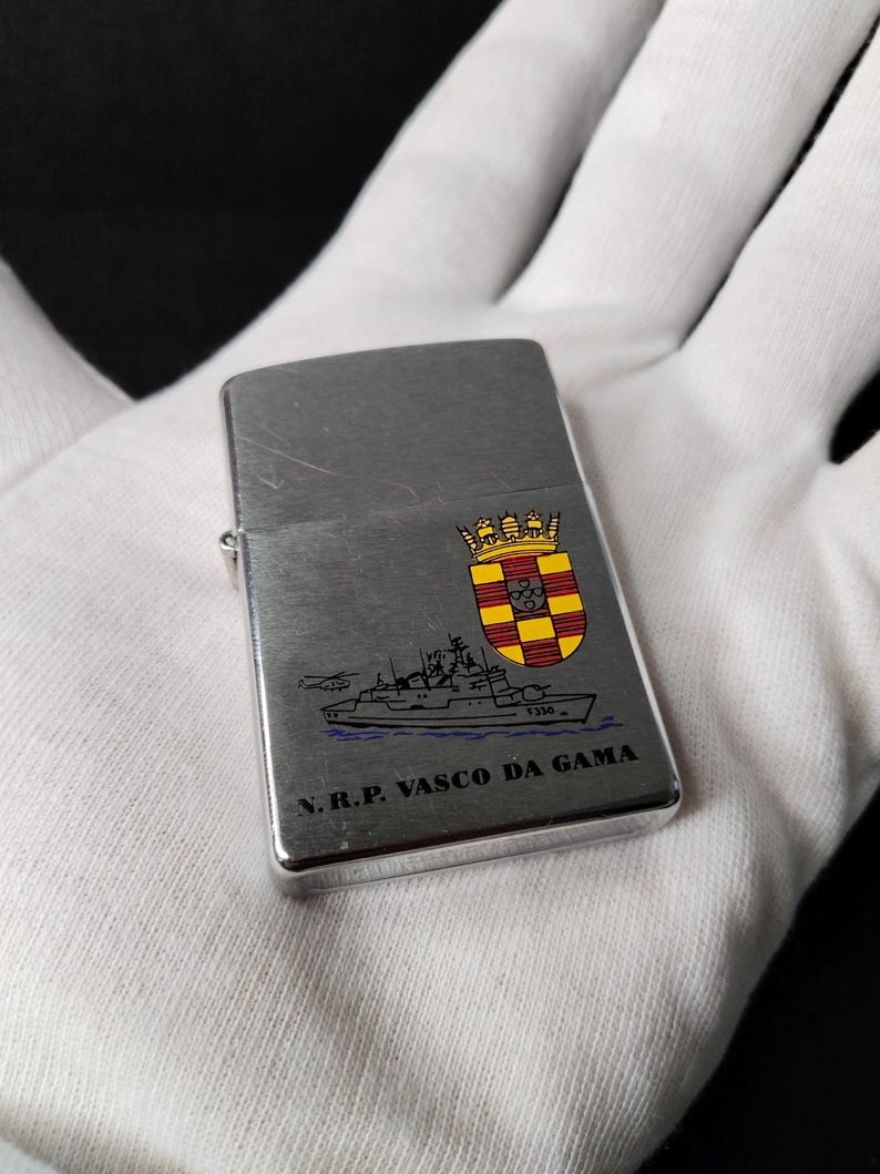 Może przedstawiać: Srebrna metalowa zapalniczka ze szczotkowanym wykończeniem. Zapalniczka posiada kolorowy emblemat i ilustrację statku z napisem "N.R.P. VASCO DA GAMA".