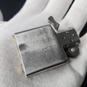 Peut inclure: Un briquet Zippo argent&eacute;, ouvert, montrant des signes d'usure. Le texte grav&eacute; "FOR BEST RESULTS USE ZIPPO FLINTS AND FUEL" est visible. Le briquet est tenu dans un gant blanc, r&eacute;v&eacute;lant son aspect usag&eacute;.