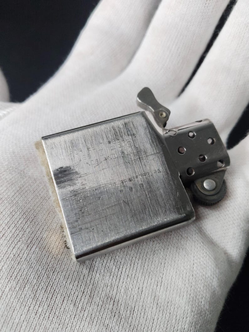 Peut inclure: Un briquet Zippo argent&eacute;, ouvert, montrant des signes d'usure. Le texte grav&eacute; "FOR BEST RESULTS USE ZIPPO FLINTS AND FUEL" est visible. Le briquet est tenu dans un gant blanc, r&eacute;v&eacute;lant son aspect usag&eacute;.