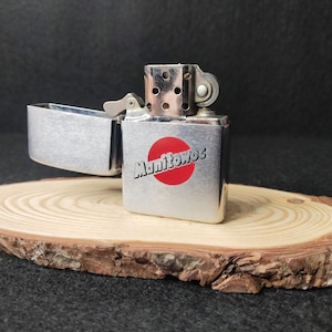 Peut inclure: Un briquet argent&eacute;, couvercle ouvert, avec le mot "Manitowoc" en blanc sur un cercle rouge. Il repose sur une petite tranche de bois ronde. Fond sombre.