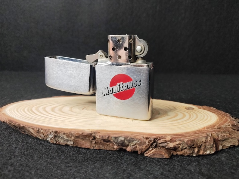 Peut inclure: Un briquet argent&eacute;, couvercle ouvert, avec le mot "Manitowoc" en blanc sur un cercle rouge. Il repose sur une petite tranche de bois ronde. Fond sombre.