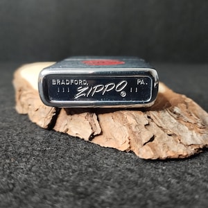 Peut inclure: Un briquet Zippo argent&eacute; avec le nom de la marque et "BRADFORD, PA." imprim&eacute; en bas. Le briquet a un embl&egrave;me rouge sur le dessus et repose sur un morceau de bois.