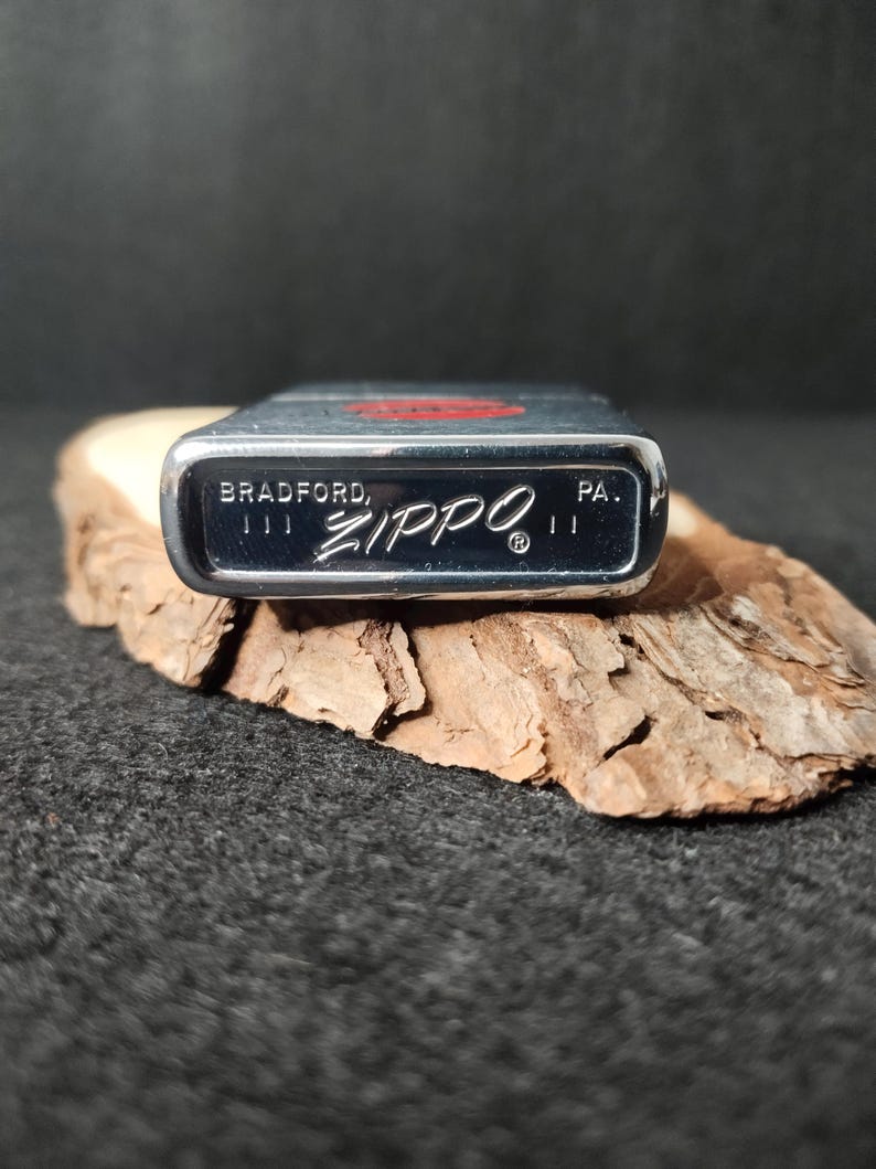 Peut inclure: Un briquet Zippo argent&eacute; avec le nom de la marque et "BRADFORD, PA." imprim&eacute; en bas. Le briquet a un embl&egrave;me rouge sur le dessus et repose sur un morceau de bois.