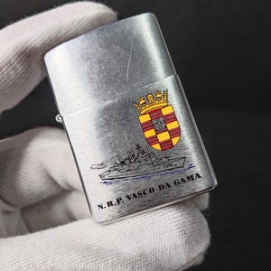 Może przedstawiać: Srebrna zapalniczka z ilustracją statku i napisem "N.R.P. VASCO DA GAMA". Zapalniczka posiada r&oacute;wnież herb z czerwonymi, ż&oacute;łtymi i złotymi akcentami. Zapalniczka jest trzymana przez dłoń w rękawicy.