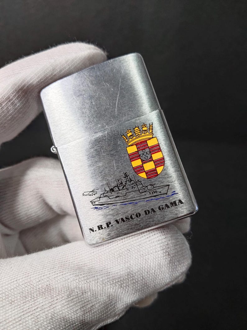 Może przedstawiać: Srebrna zapalniczka z ilustracją statku i napisem "N.R.P. VASCO DA GAMA". Zapalniczka posiada r&oacute;wnież herb z czerwonymi, ż&oacute;łtymi i złotymi akcentami. Zapalniczka jest trzymana przez dłoń w rękawicy.