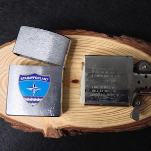 Może przedstawiać: Srebrna zapalniczka z otwartą pokrywą, prezentująca niebiesko-biały emblemat z napisem "STANAVFORLANT". Zapalniczka leży na drewnianej powierzchni. Tekst wewnątrz brzmi "KEEP AWAY FROM CHILDREN" i "LIGHTER DOES NOT SELF EXTINGUISH".